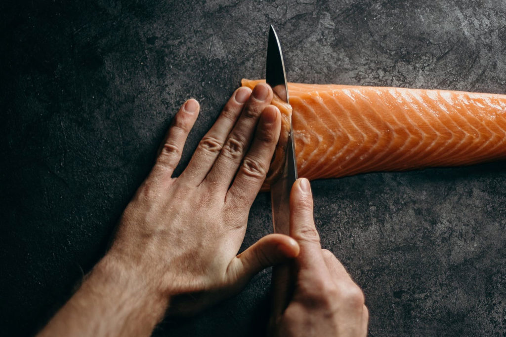 a close up photo of a person's hands using a knife to cut a fillet of salmon on a piece of slate