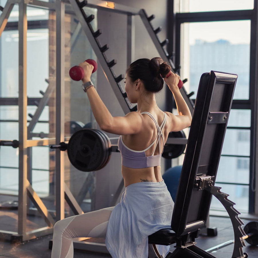 overhead-press-woman-on-bench-in-gym a woman in a gym sitting upright on a bench doing an overhead press with free weights