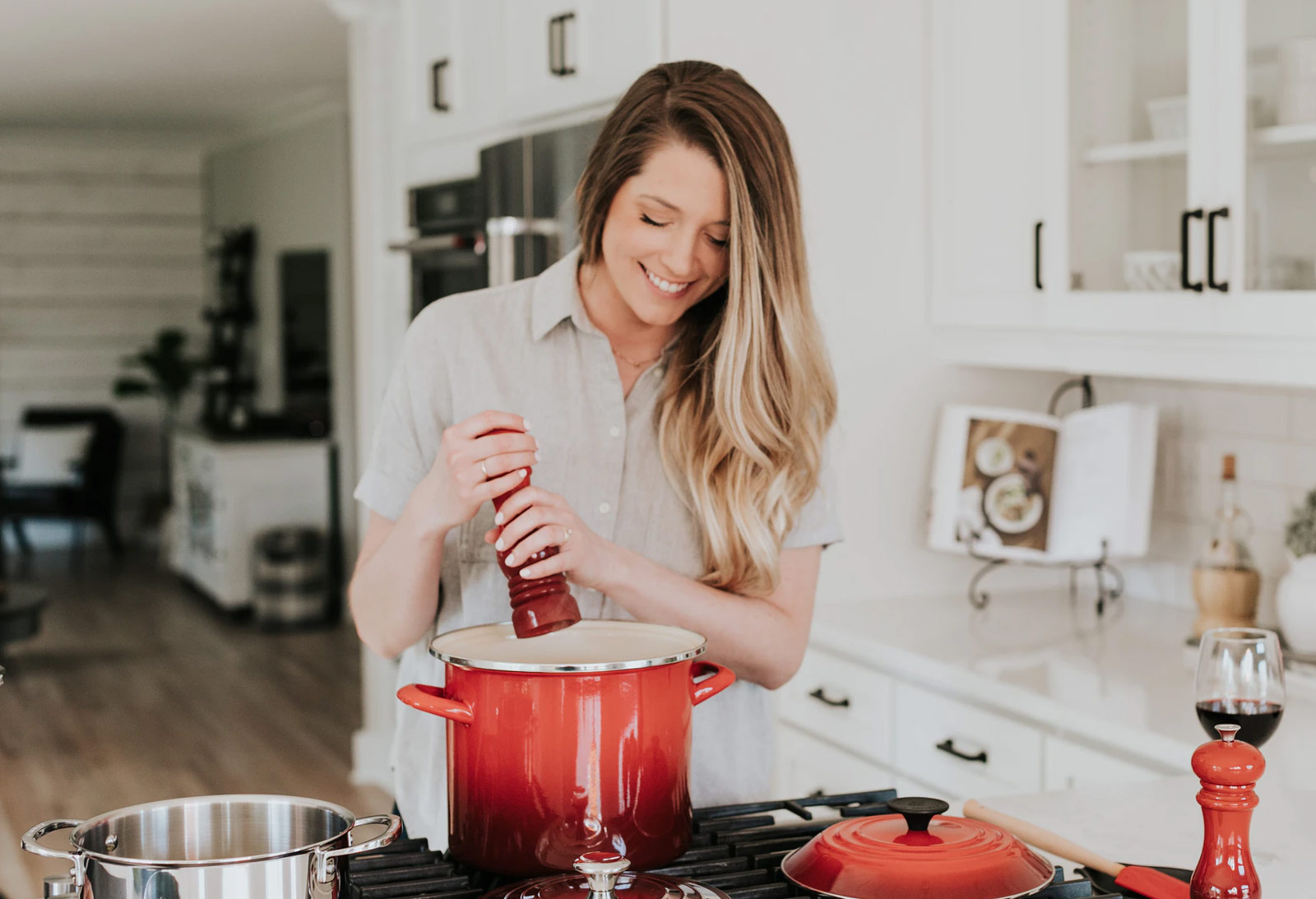 a woman smiling in the kitchen while grinding pepper into a large red pot