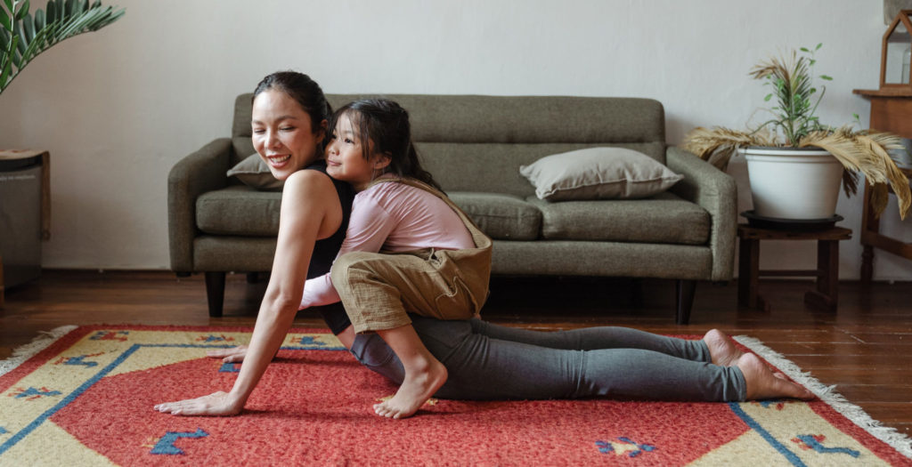 a woman doing yoga on a rug in her home while her young daughter hugs her