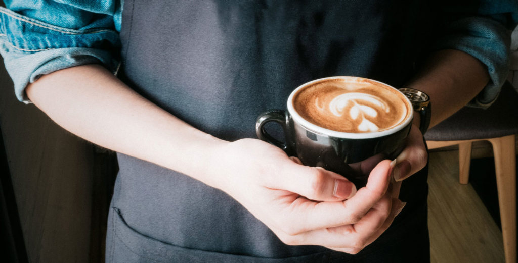 a barista wearing a denim jacket and apron holding a cup of fresh coffee