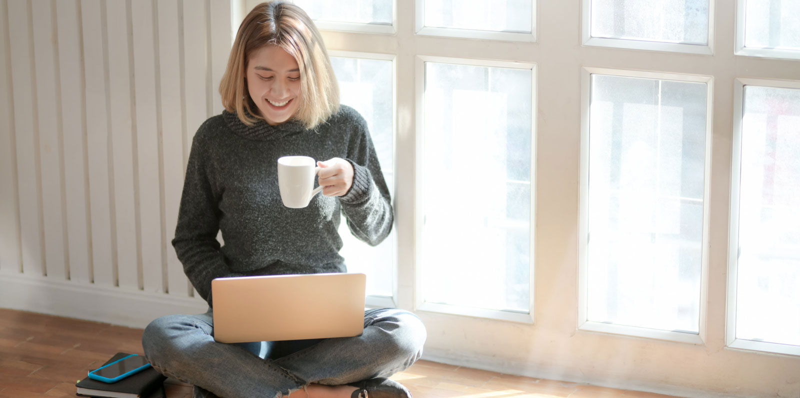 a young woman smiling while holding a cup of coffee and sitting cross legged on the floor with a laptop