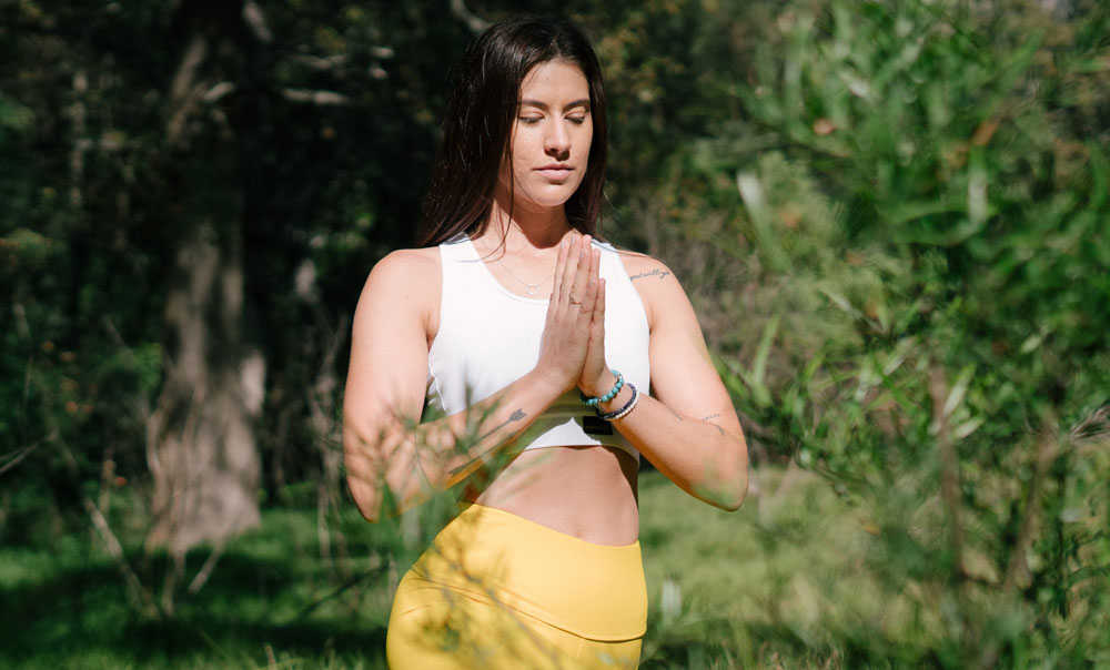 a woman wearing a white sports bra and yellow yoga pants doing meditation in nature