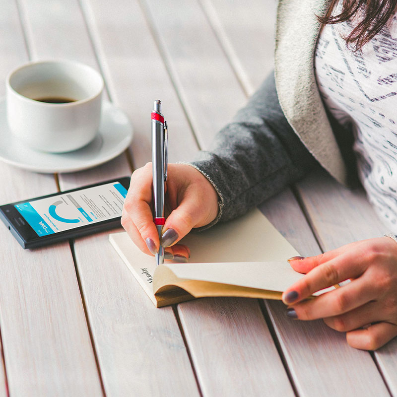 woman writing into a notebook on a wooden table with a phone and coffee