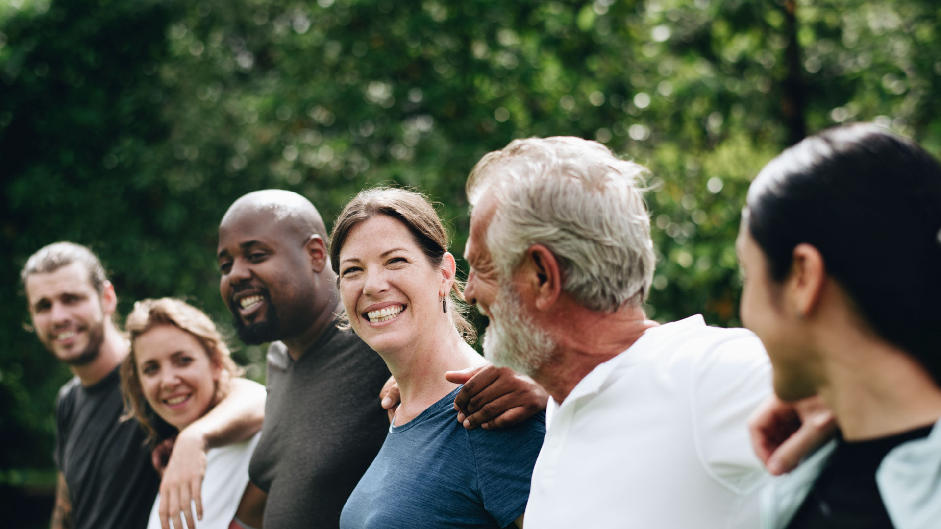 a smiling group of people standing in a line outdoors with arms around each other's shoulders