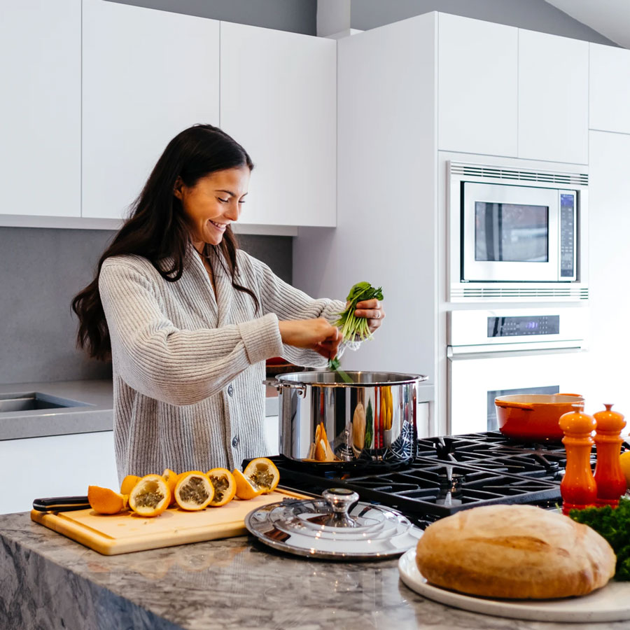 a smiling woman wearing a striped gown in the kitchen dropping leafy vegetables into a pot