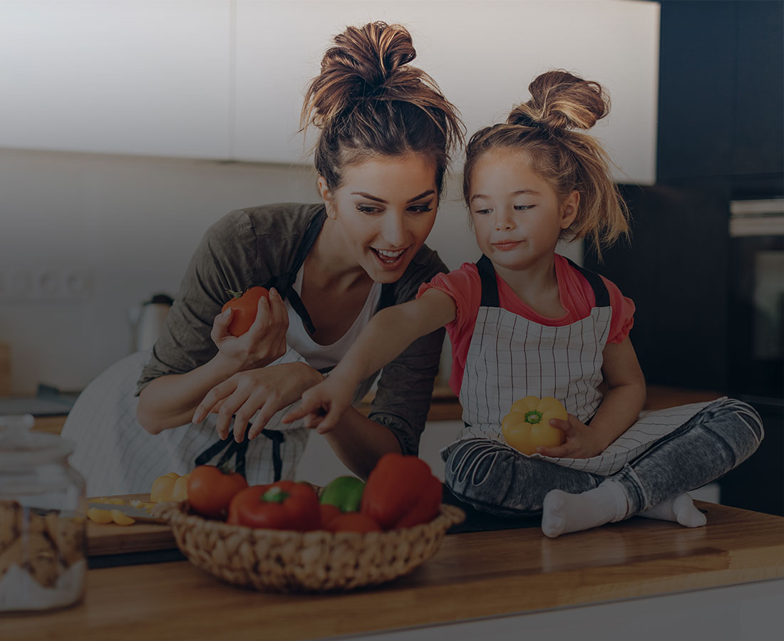 a young girl sitting on a table picking out vegetables with her mom