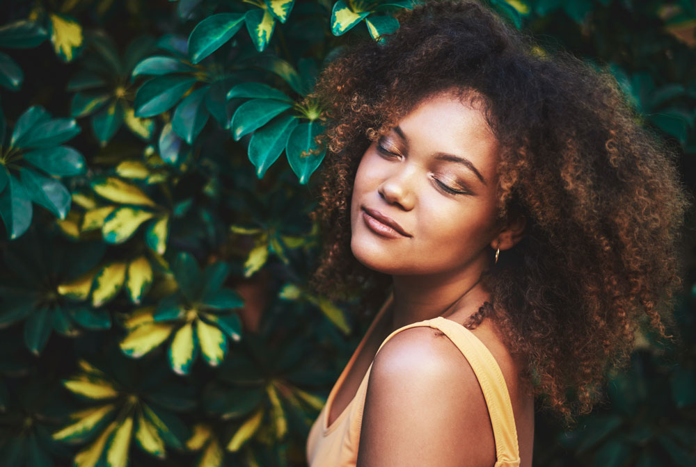 a woman with curly hair and glowing skin standing in front of green leaves